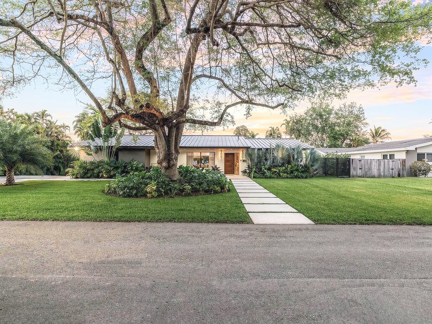 Palmetto Bay spec home at dusk beneath a mature oak, with a wood-grain entrance door at the end of the front walk